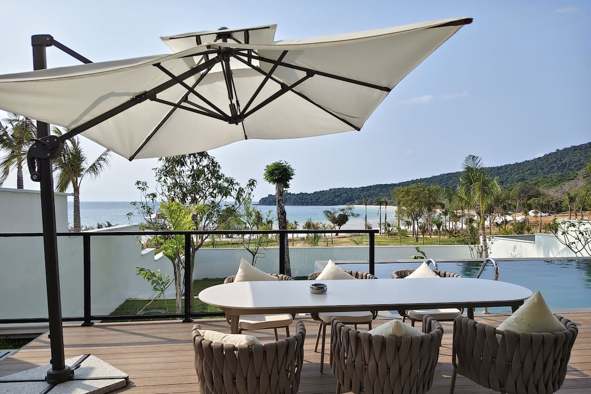An outdoor seating area with a table and chairs, a large umbrella, and a view of the sea and hills.