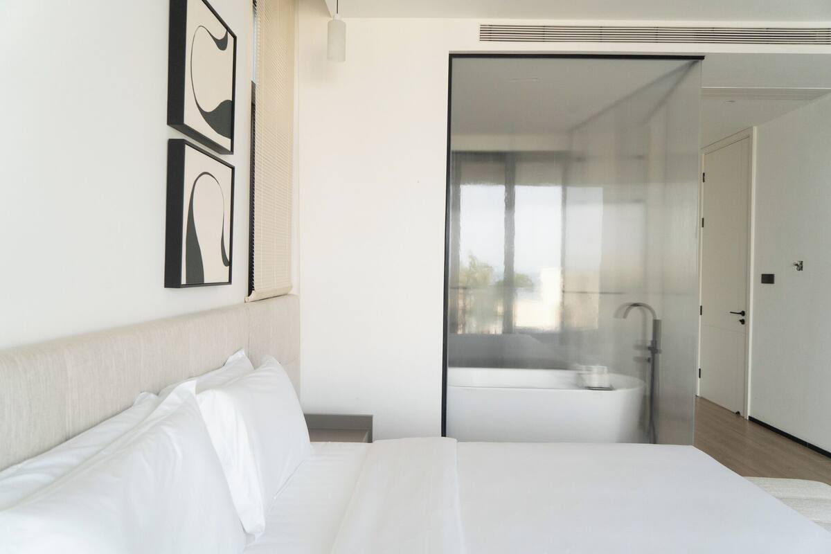 A modern hotel room with a large bed, a glass partition, and a visible bathroom with a bathtub.