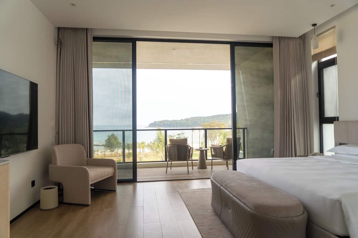 A modern hotel room with a large bed, a seating area with two chairs, a TV, and a balcony with a view of the sea.