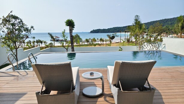 A poolside area with two wicker chairs and a small table, overlooking a beach and ocean.