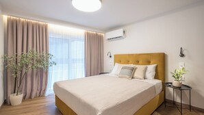 Quarto de hotel moderno com uma cama grande, uma planta em vaso, uma mesa de cabeceira e uma janela com cortinas.