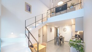 Um interior moderno com uma escada que leva a um mezanino, uma área de jantar com mesa e cadeiras, e uma cozinha com bancada e eletrodomésticos.