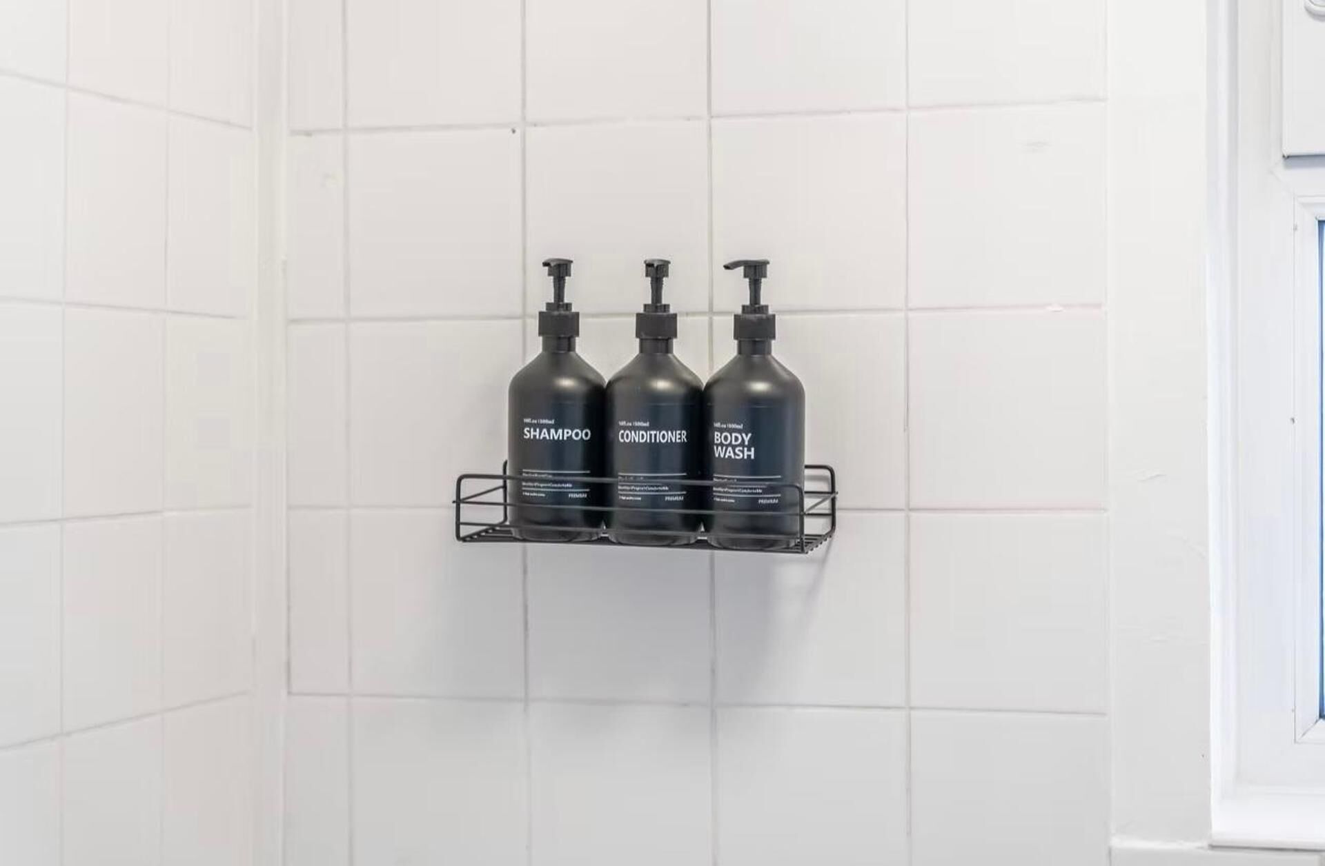 A bathroom with three black bottles of shampoo, conditioner, and body wash on a metal shelf against a white tiled wall.