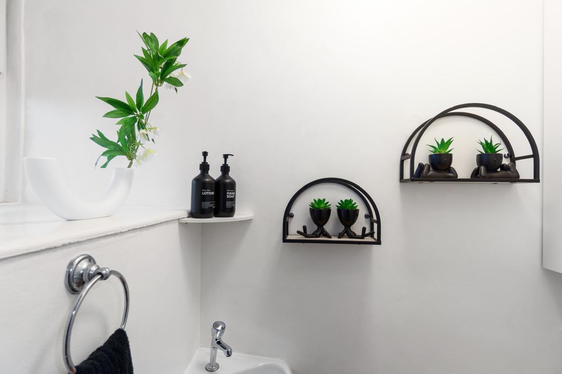 A bathroom with a white sink, a black towel ring, a white bowl, two black lotion bottles, and two potted plants on shelves.