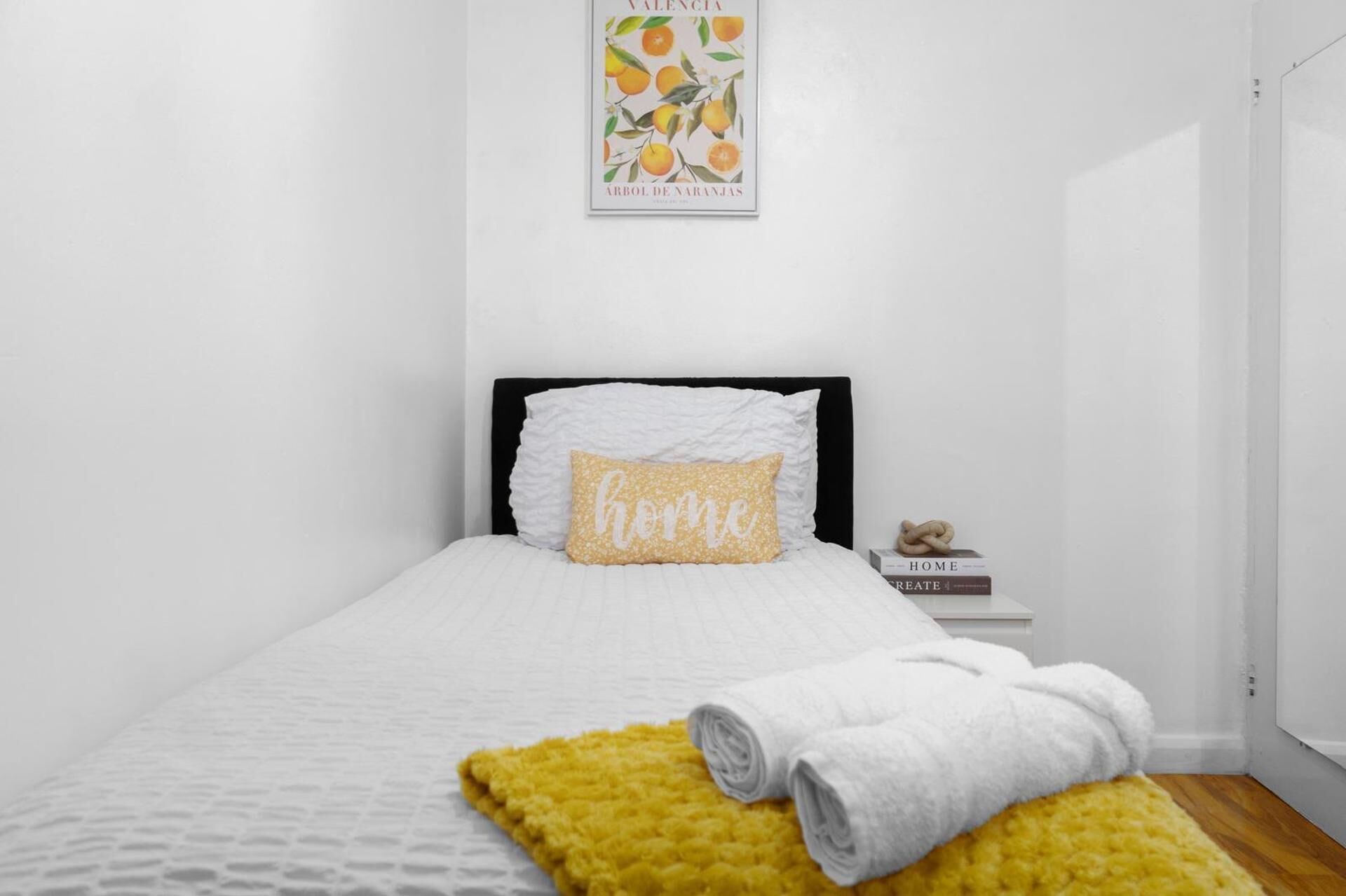 A neatly made bed with a white duvet and a yellow throw blanket, a pillow with the word 'home,' and a framed picture of oranges on the wall.