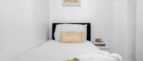 A neatly made bed with a white duvet and a yellow throw blanket, a pillow with the word 'home,' and a framed picture of oranges on the wall.