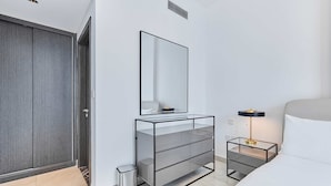 A modern hotel room with a large mirror, a bedside table with a lamp, a glass storage unit, and a trash bin. - Brand New 1B With Burj Khalifa View in Business Bay (Dubai)