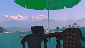Una mesa con una computadora portátil, un vaso de agua y un paraguas verde con la marca 'TUBORG', con vistas a un lago y montañas al fondo.