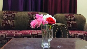 Un florero de vidrio transparente con flores rosadas y rojas sobre una mesa de madera en una habitación con sofás estampados y una cortina oscura al fondo.