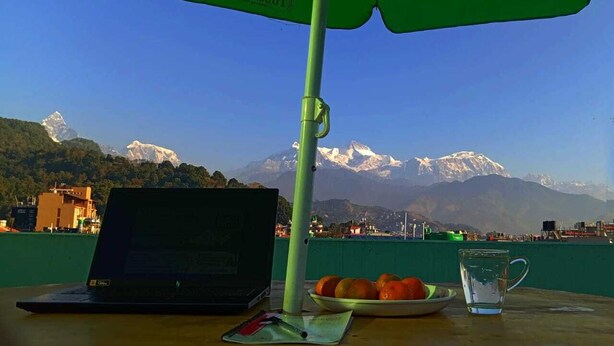 Una computadora portátil, un plato de naranjas y un vaso de agua sobre una mesa con un paraguas verde, con vistas a un paisaje montañoso.