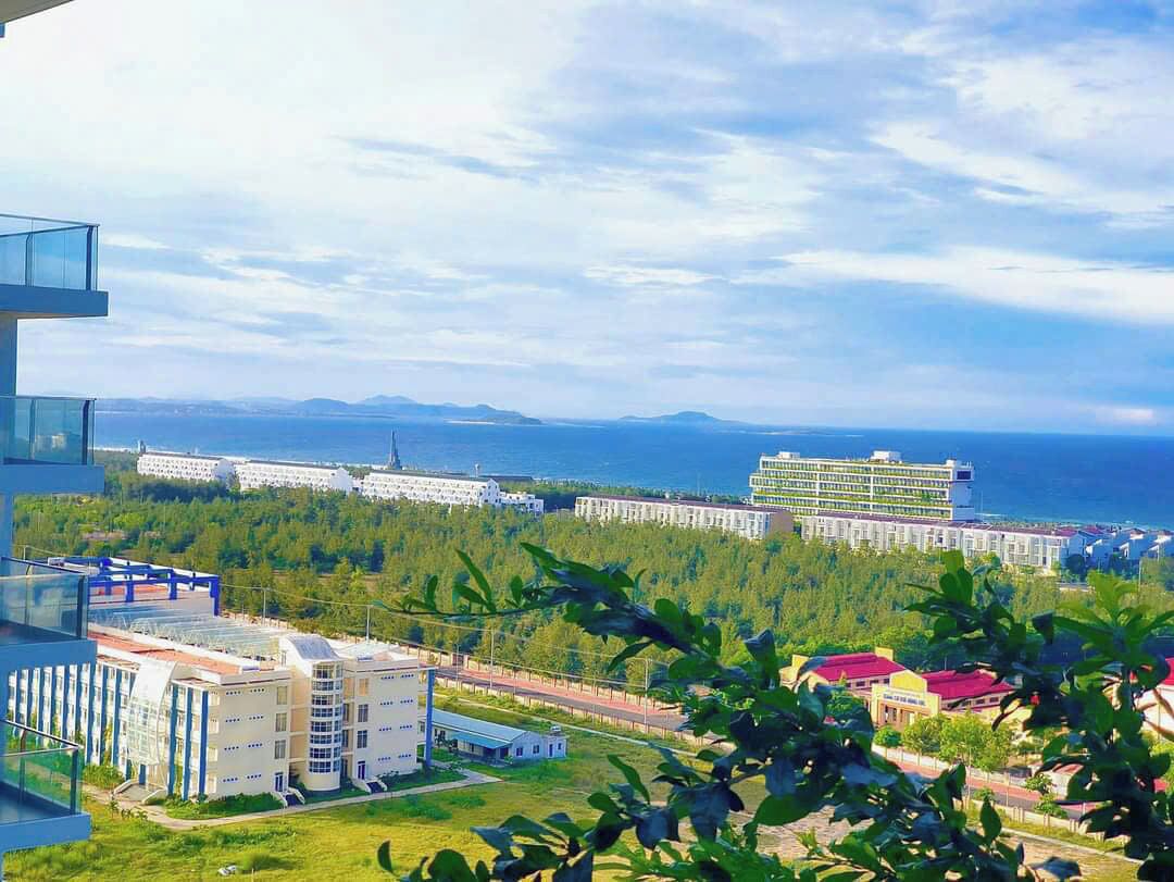A modern hotel with a glass facade, located near a coastline with a view of the sea and distant mountains.