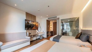 A modern hotel room with two beds, a sofa, a TV, and a bathroom visible through a glass door.