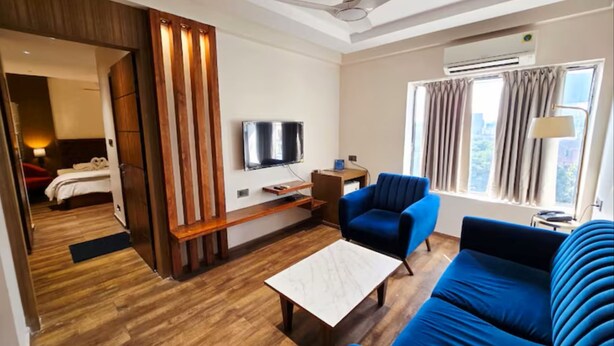 Una camera d'albergo con pavimento in legno, una ventola a soffitto, una TV, un divano, un tavolino da caffè e una finestra con tende.