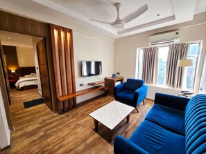 Habitación de hotel con piso de madera, ventilador de techo, televisor, sofá, mesa de centro y ventana con cortinas.