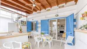 Un patio iluminado por el sol con una mesa redonda, sillas blancas y contraventanas azules en las paredes.