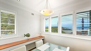 A dining area with a glass table, white chairs, a wooden bench, and a view of the sea through the windows. - RAG25 - Balmoral Beach Lifesaver – Executive Listing (Mosman)