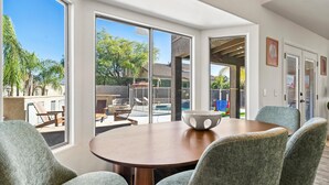 Ein Essbereich mit einem runden Tisch, vier grauen Stühlen und einer Schüssel. Große Fenster bieten Blick auf einen Garten mit Pool, Stühlen und einer Spielanlage.