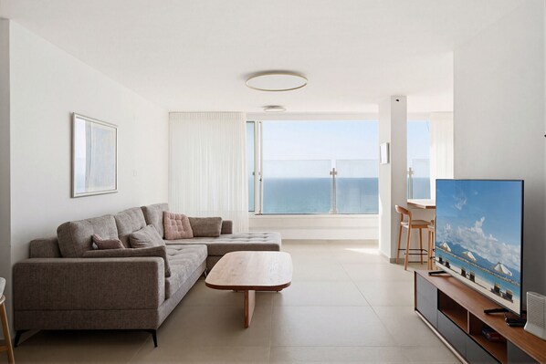 A modern living room with a sofa, a coffee table, a TV, and a view of the sea through a large window. - MIKLAT Family Friendly & Stunning Sea View by FeelHome (Netanya)