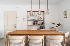 A dining area with a wooden table and six chairs, a bookshelf with decorative items, and pendant lights. - MIKLAT Family Friendly & Stunning Sea View by FeelHome (Netanya)