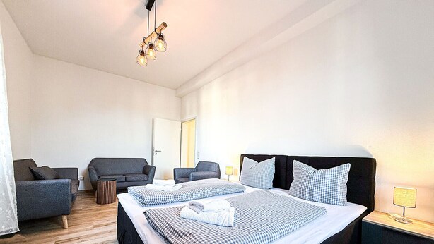 Quarto de hotel com uma cama grande, duas poltronas, uma mesa pequena e um candeeiro suspenso.