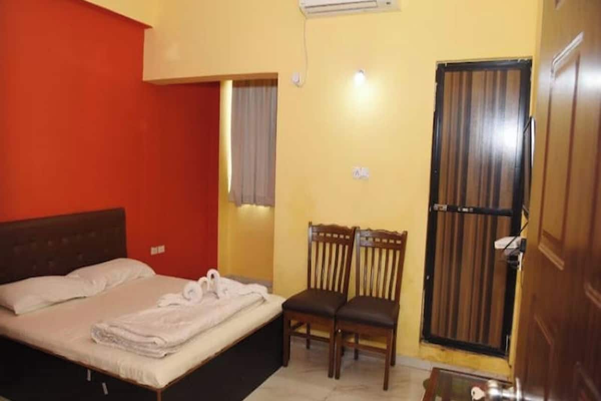 Un dormitorio con cama, dos sillas, una mesita, aire acondicionado y ventana con cortinas.
