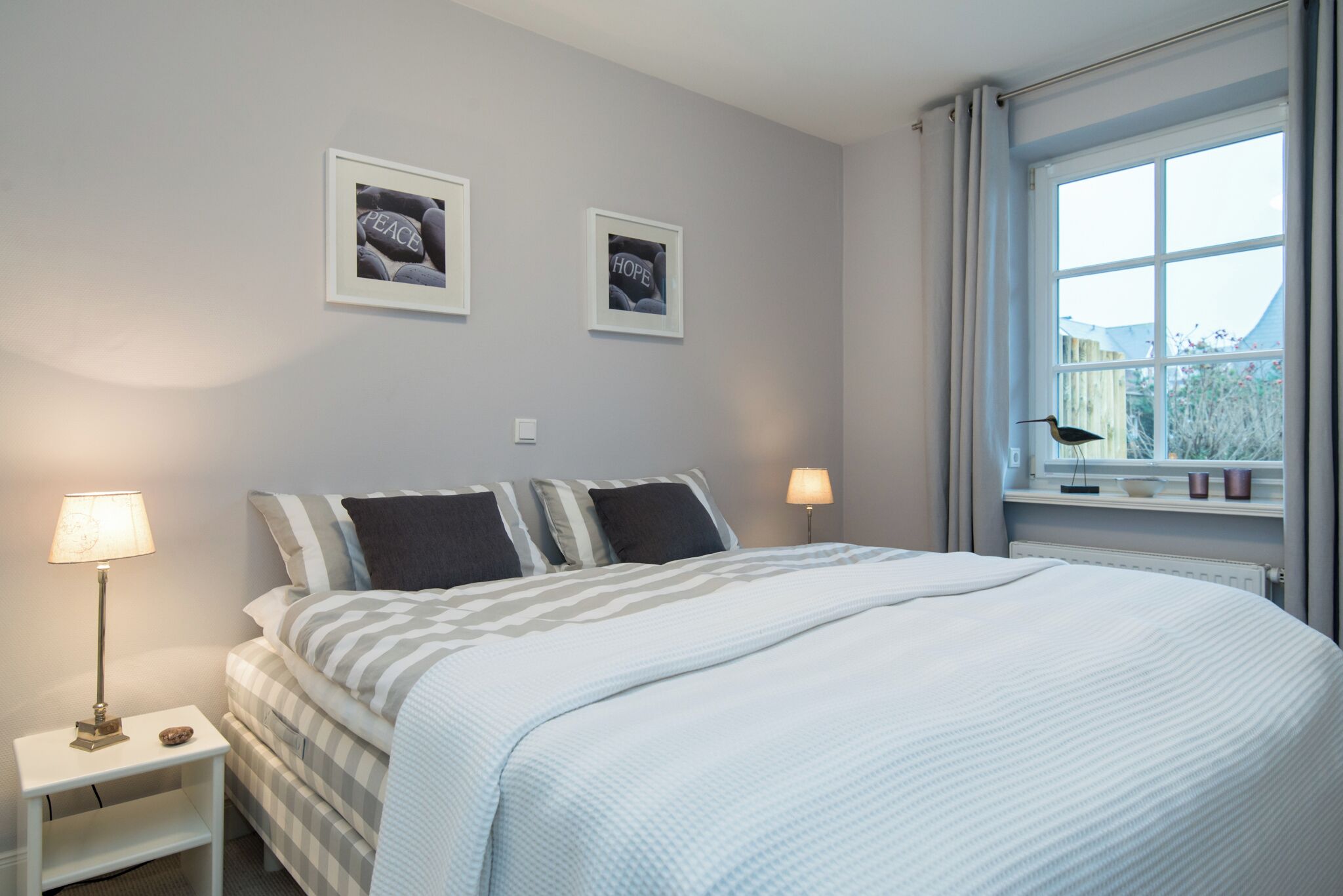 A bedroom with a bed, two bedside tables, a window, and two framed pictures on the wall.