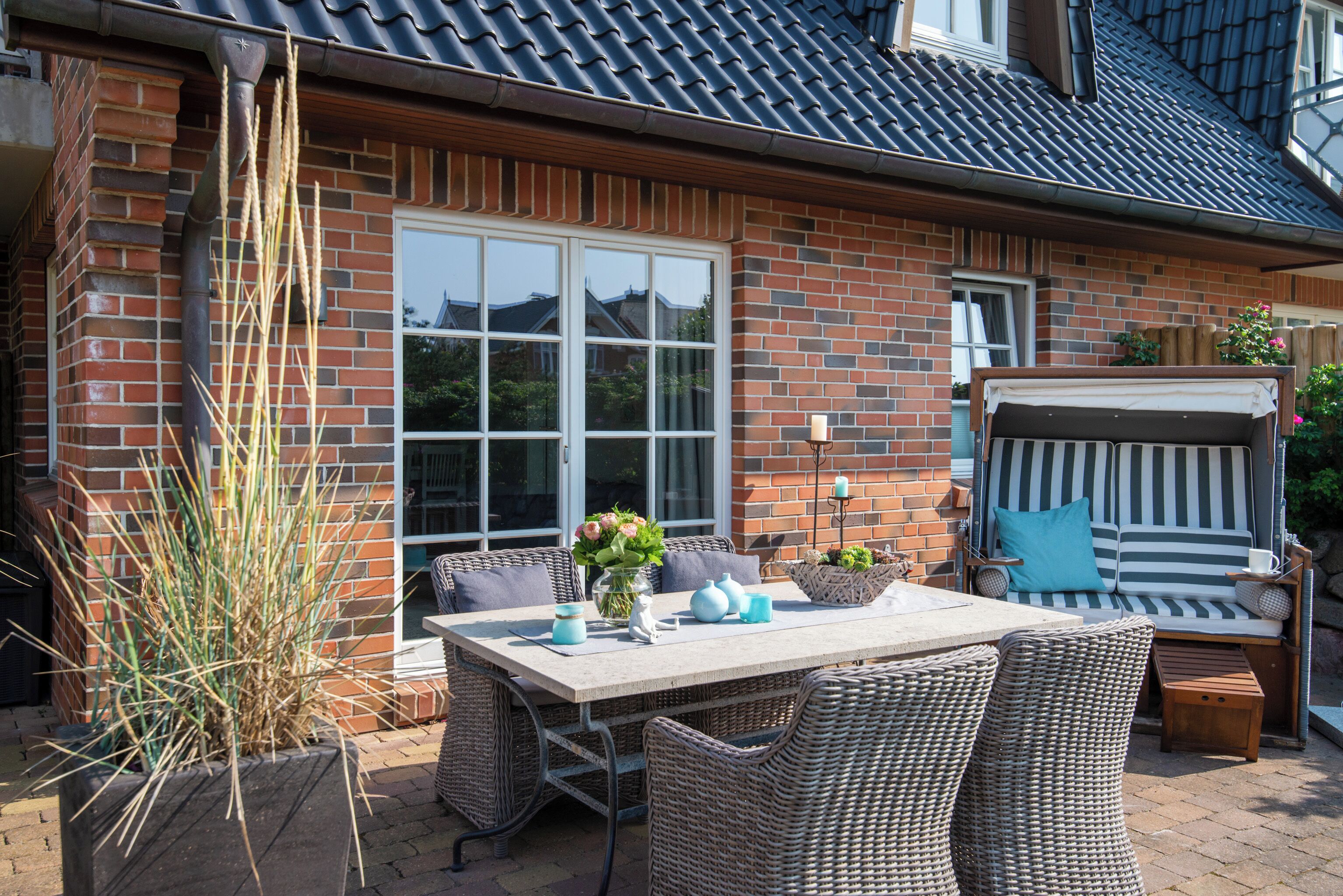 A patio with a dining table, wicker chairs, a cushioned outdoor seating area, and a brick house with large windows.