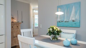A dining area with a white table, chairs, and a vase of flowers. There is a painting of sailboats on the wall. - Exclusive Anker Mal Sylt vacation apartment with terrace and garden on the sandy beach (Sylt)