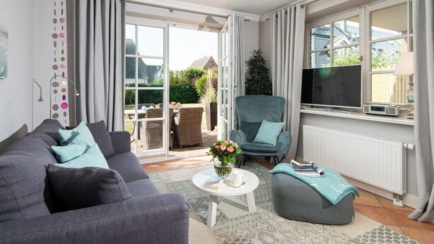 A modern living room with a grey sofa, a teal armchair, and a white coffee table. There is a large window with curtains, a TV, and a radiator. - Exclusive Anker Mal Sylt vacation apartment with terrace and garden on the sandy beach (Sylt)