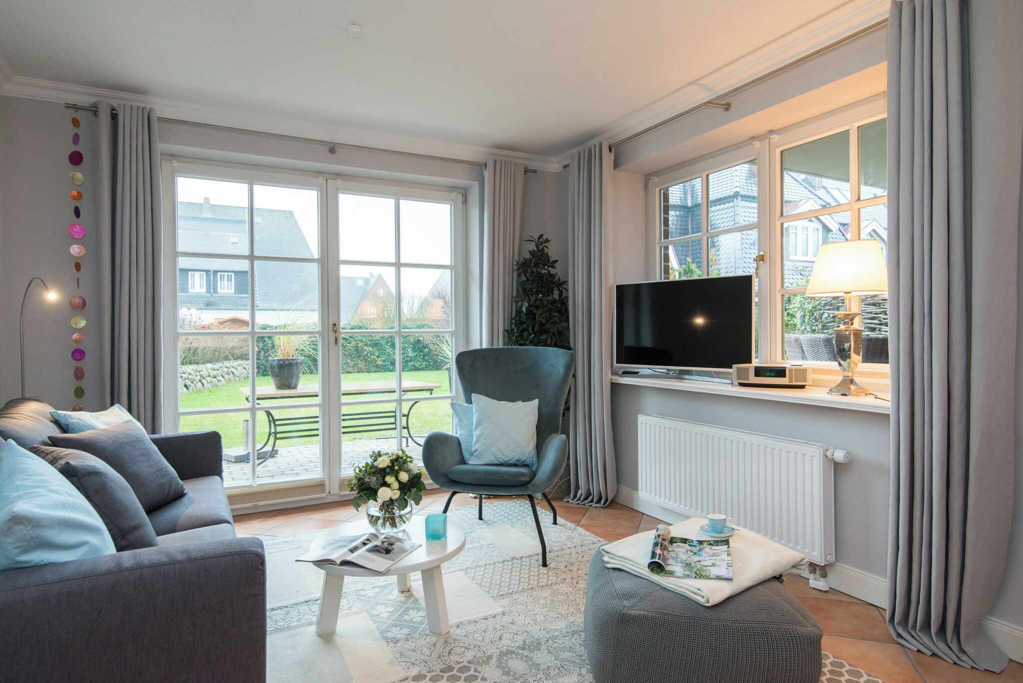 Ett vardagsrum med en grå soffa, en blå fåtölj, ett soffbord, en tv och ett fönster med gardiner.