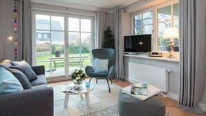 A living room with a grey sofa, a blue armchair, a coffee table, a TV, and a window with curtains. - Exclusive Anker Mal Sylt vacation apartment with terrace and garden on the sandy beach (Sylt)