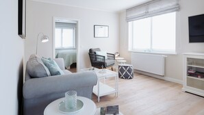 Uma sala de estar moderna com um sofá cinzento, uma pequena mesa de centro branca, uma poltrona de vime preta e um radiador branco.