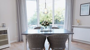 Uma sala de jantar com uma mesa, duas cadeiras de vime, um vaso com flores e vista para o verde lá fora.