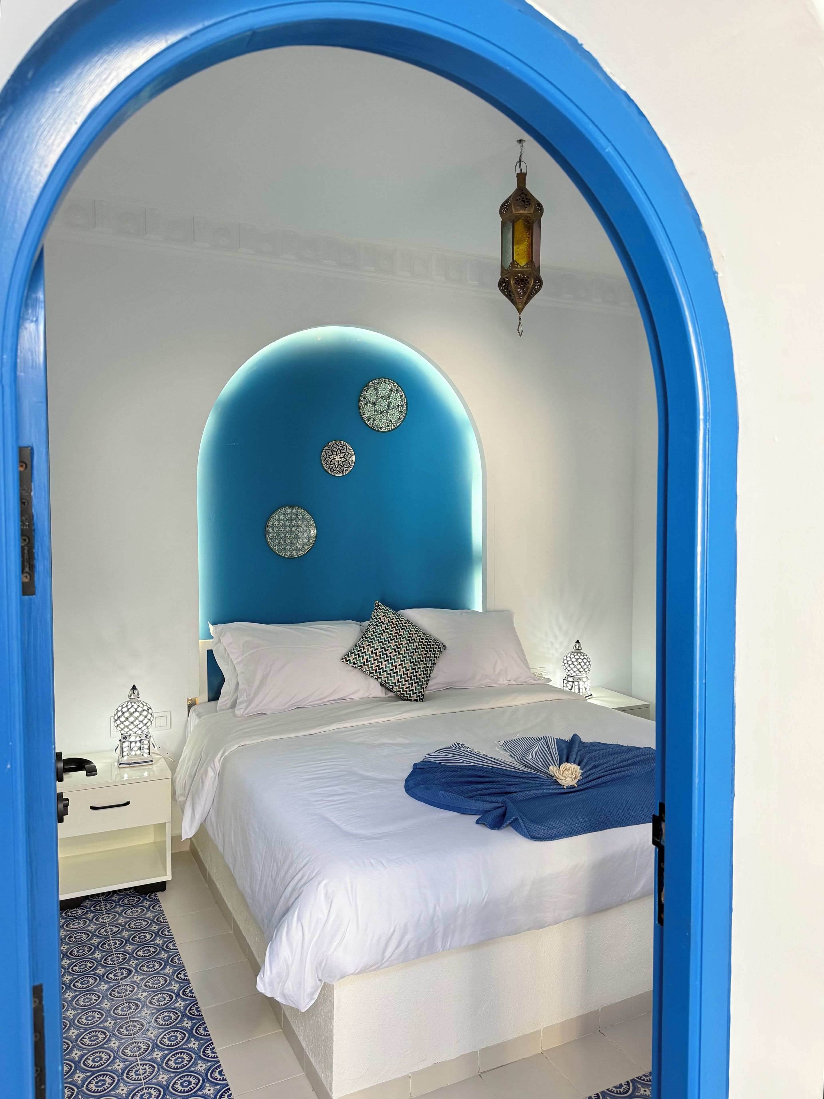 A bedroom with a blue archway, a bed with white bedding, patterned pillows, and a decorative hanging light.