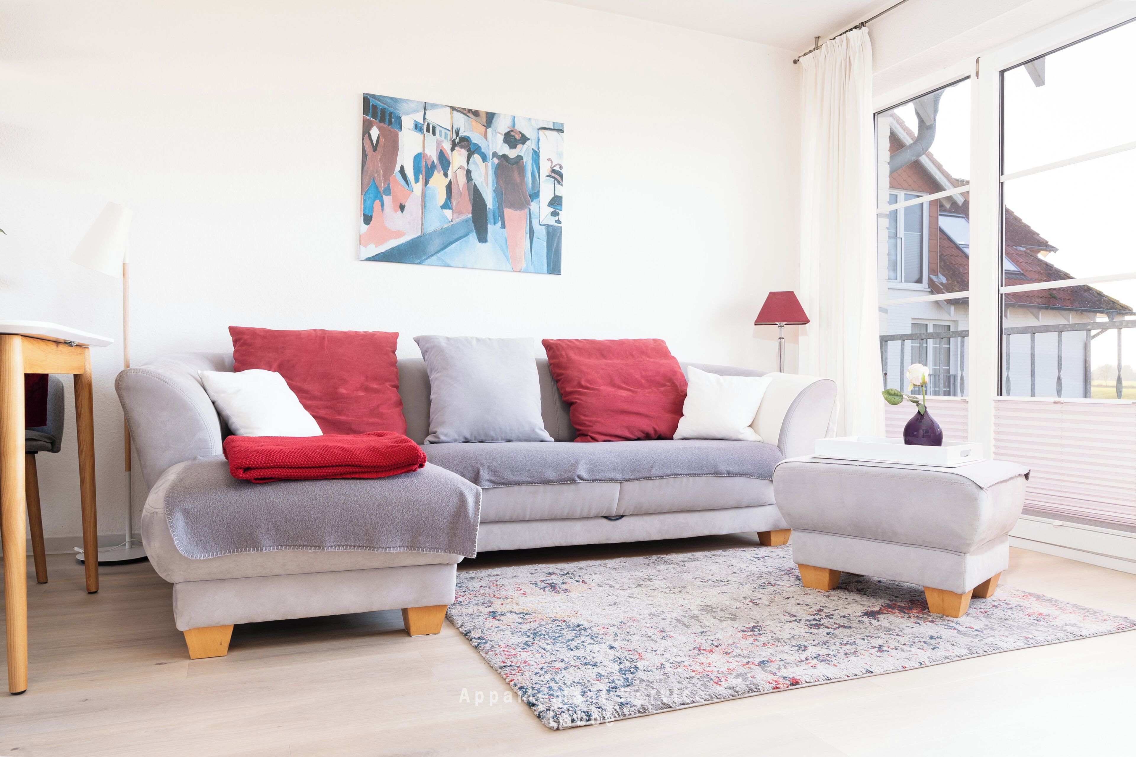 Ein modernes Wohnzimmer mit einem grauen Sofa, roten und weißen Kissen, einem kleinen Beistelltisch und einem großen Fenster.