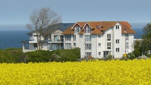 Un edificio grande de color blanco con techo rojo, múltiples ventanas y balcones, ubicado cerca de un campo de flores amarillas y con vistas al mar.