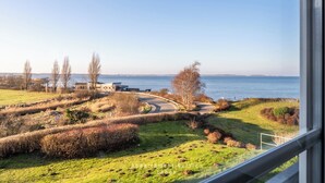 Vista desde una ventana sobre un paisaje costero con una carretera serpenteante, campos verdes y una masa de agua.