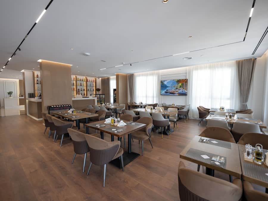 A modern restaurant with multiple dining tables and chairs, a bar area with bottles, and a large window with a painting.