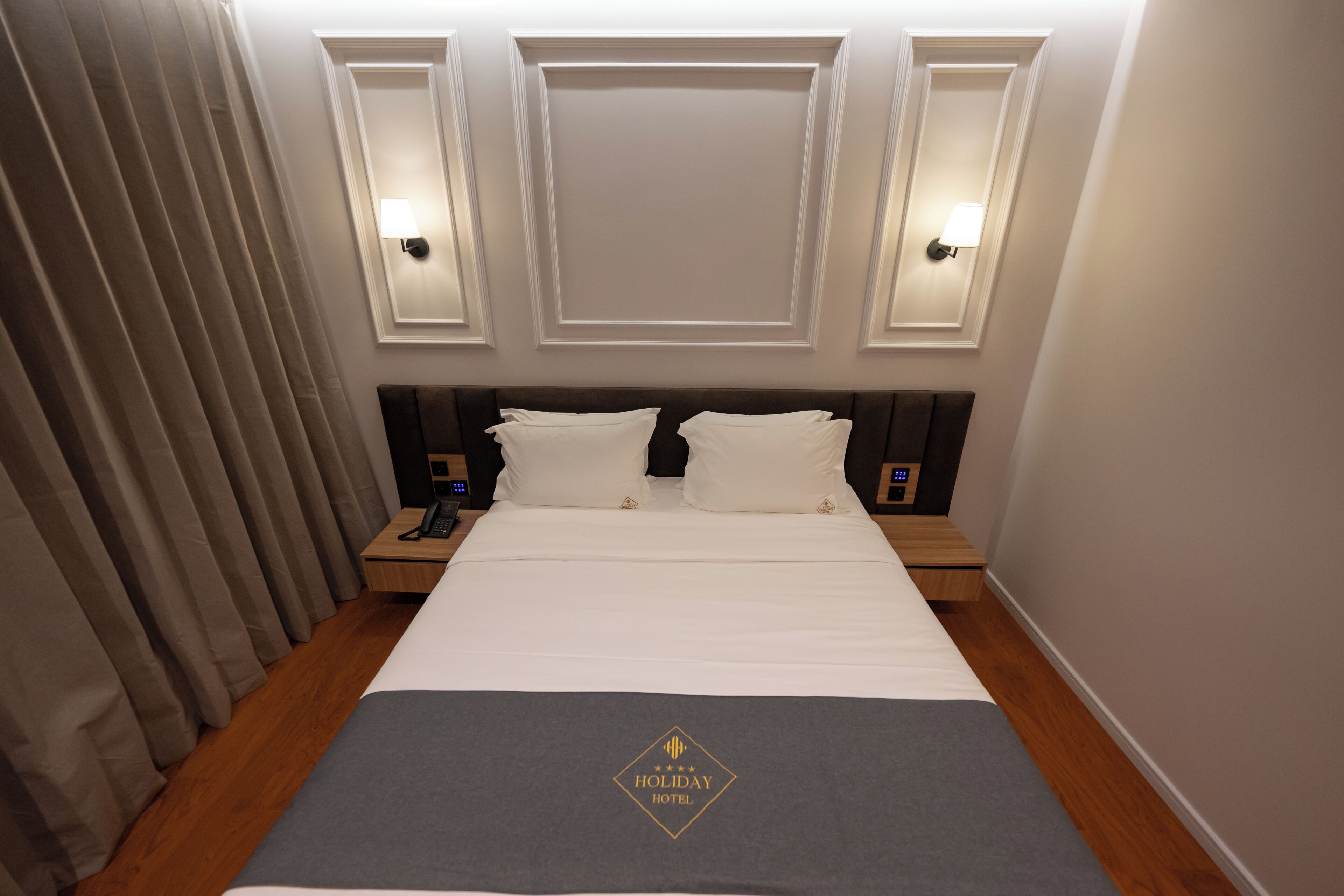 Una habitación de hotel con cama, mesitas de noche, teléfono y lámpara de pared.