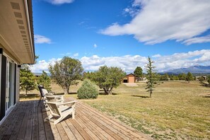Una terraza de madera con sillas, cielo despejado y un campo con césped, árboles y una montaña al fondo.