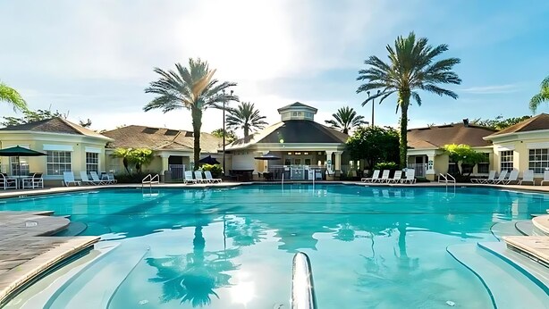 A swimming pool with lounge chairs and umbrellas, surrounded by palm trees and buildings with a Mediterranean architectural style.