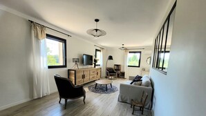 Amplia sala de estar con piso de madera, un sillón de cuero negro, un sofá blanco, un mueble de madera con un televisor y una estantería.