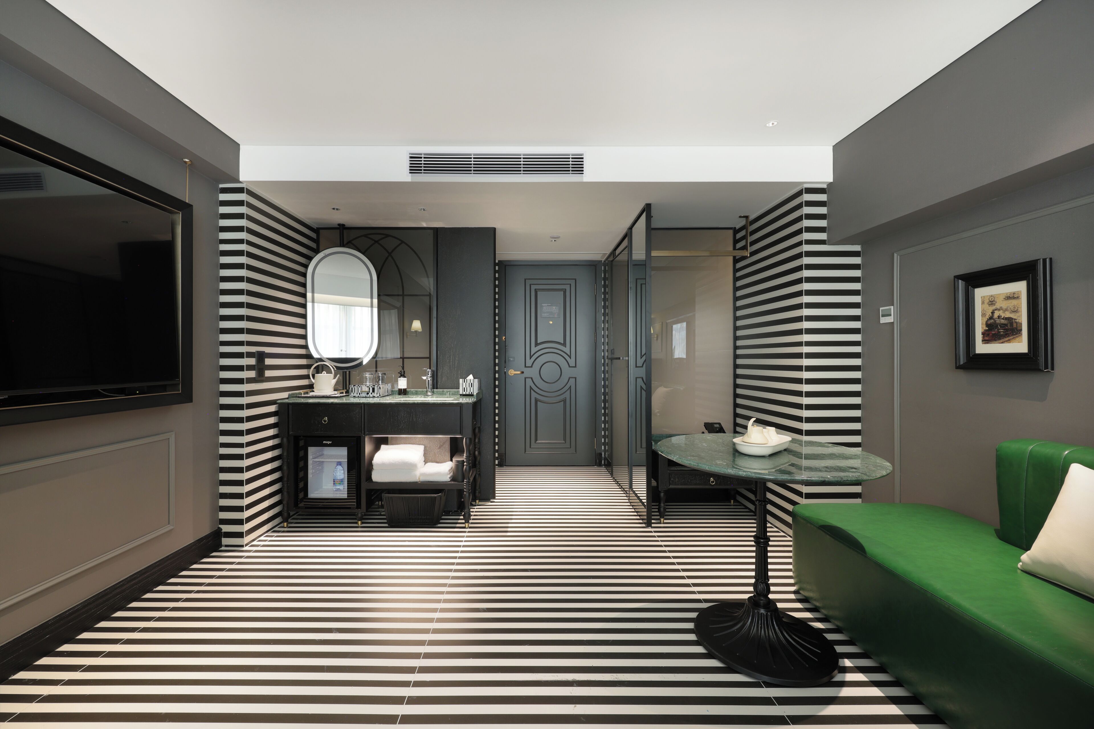 Habitación de hotel con piso a rayas, un sofá verde, una mesita, un espejo, un televisor y una puerta con detalles decorativos.