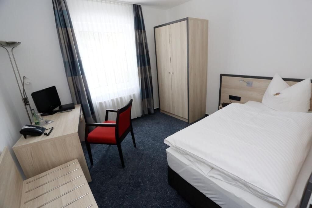 Ein Hotelzimmer mit Bett, Schreibtisch mit Computer, Stuhl, Kleiderschrank und Fenster mit Vorhängen.
