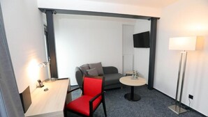 Ein kleines Hotelzimmer mit Schreibtisch, einem roten Sessel, einem grauen Schlafsofa, einem runden Tisch, einer Stehlampe und einem an der Wand befestigten Fernseher.