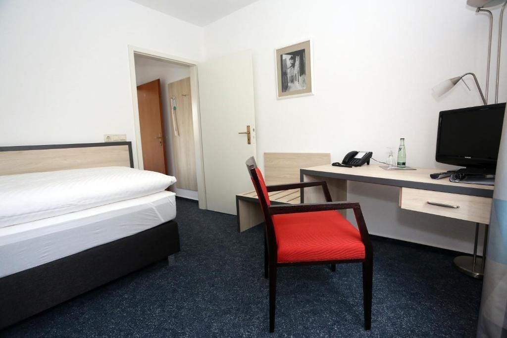 Ein Hotelzimmer mit einem Bett, einem Schreibtisch mit Stuhl, einem Telefon, einem Fernseher und einer Tür zu einem weiteren Raum.