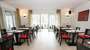 Ein Hotelrestaurant mit mehreren Tischen und Stühlen, großen Fenstern und Blick ins Freie.