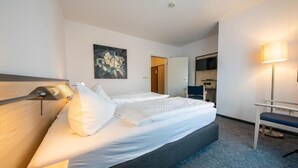 Ein Hotelzimmer mit einem großen Bett, einem Schreibtisch und einem Sessel. Ein Gemälde hängt an der Wand, und eine Tür führt zu einer Küchenzeile.