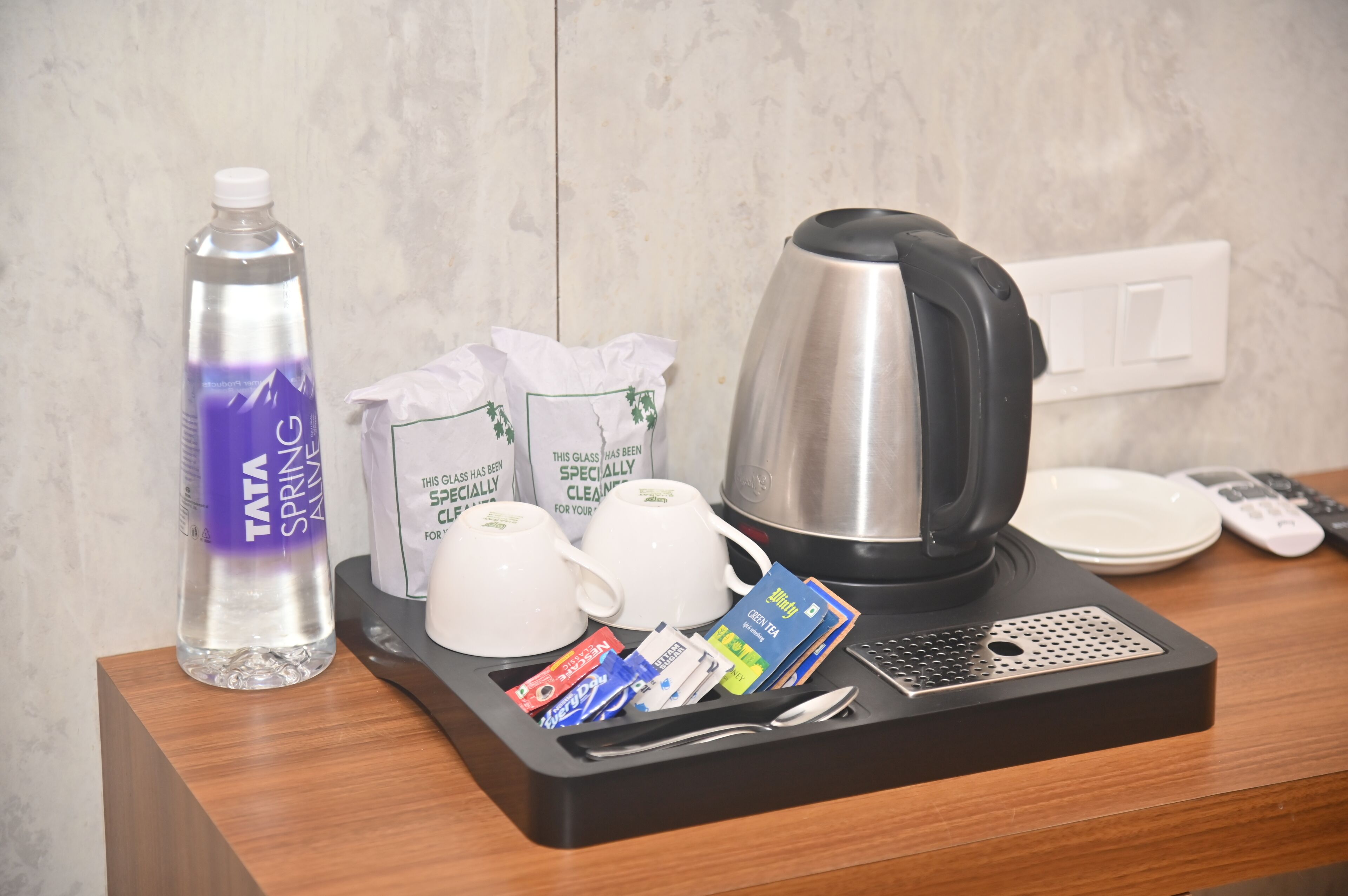 A hotel room's breakfast tray with a TATA SPRING water bottle, two white coffee cups, a black electric kettle, a tray with tea bags, a remote control, and a spoon.