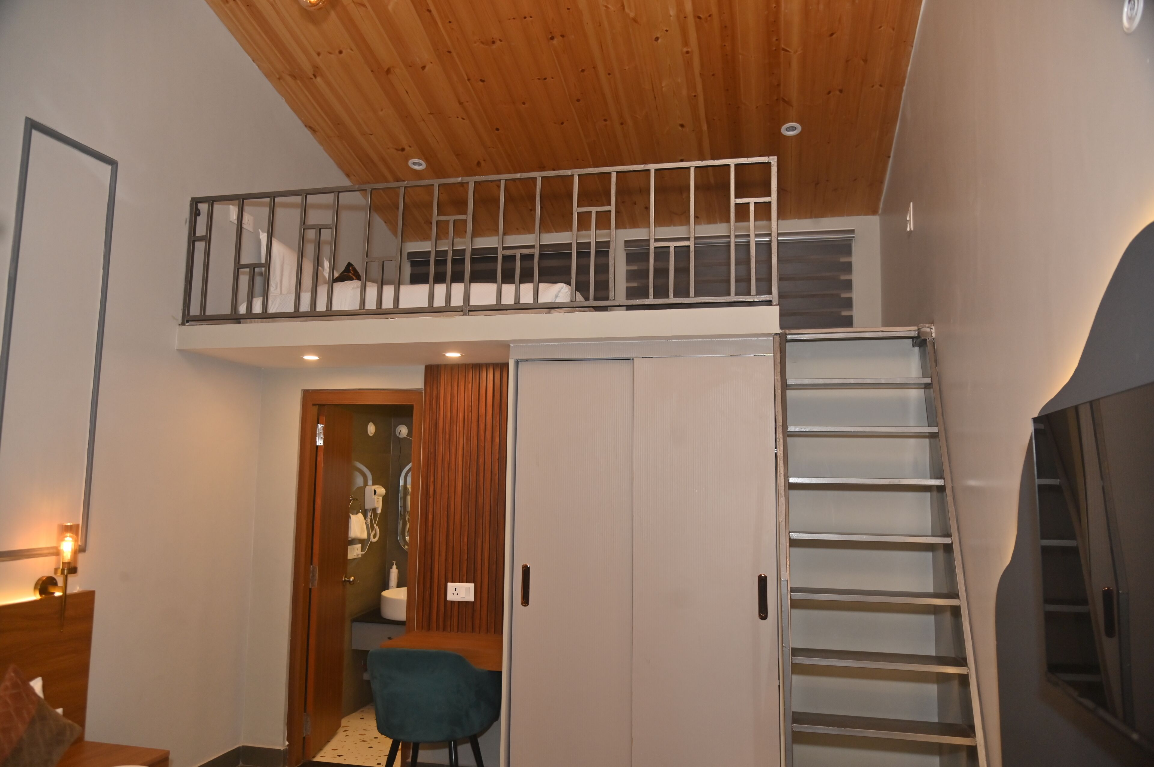 Una habitación tipo loft con techo de madera, un entresuelo con barandilla de metal, una escalera y un baño con puerta de madera.